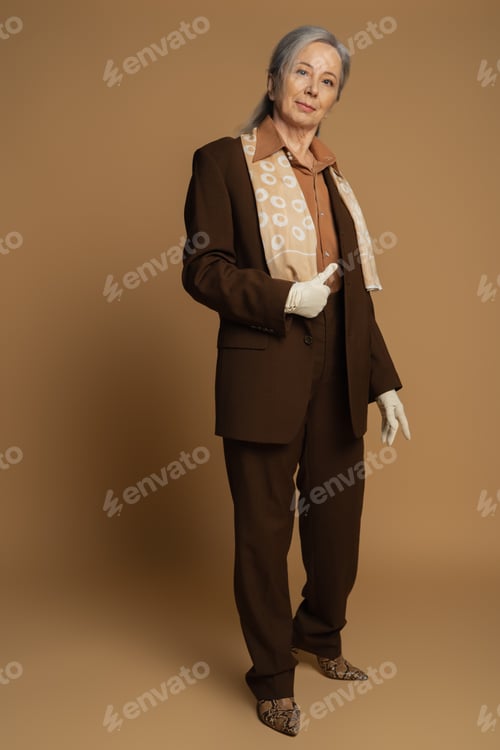 Preview: full length of senior woman in brown formal wear and white gloves showing like sign on beige
