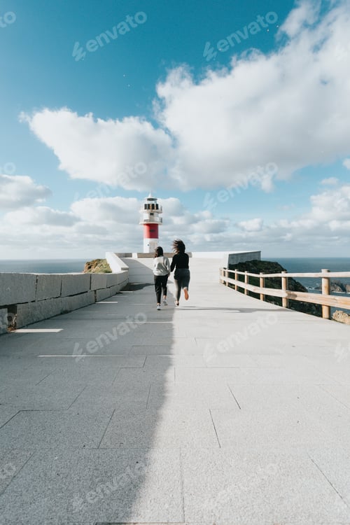 Vista previa: Dos mujeres jóvenes corriendo hacia un faro rojo y blanco, inspiración pareja y conceptos de libertad