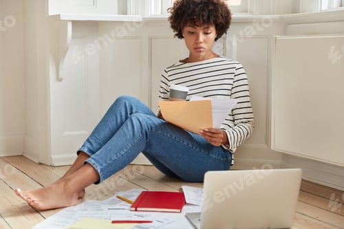 Preview: Indoor shot of pleasant looking black businesswoman has paper work at home, dressed in casul striped