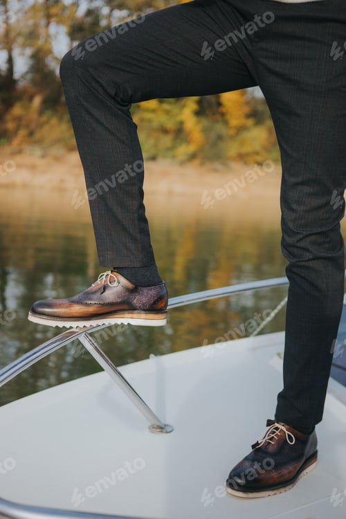 Preview: A stylish man in textured black suit pants and dark brogue shoes, standing on a boat