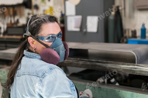 Preview: Woman wearing safety glasses and dust mask standing in metal workshop, looking at camera.