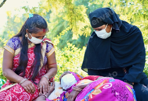 Preview: Closeup of three Indian women with one of them lying down, all wearing facemask during daylight