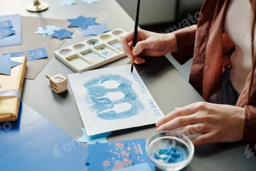 Preview: Hands of young woman with paintbrush writing down Happy Hanukkah