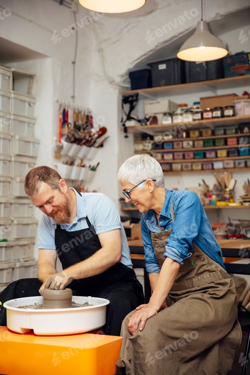 Preview: Senior woman spinning clay on a wheel with teacher at pottery class