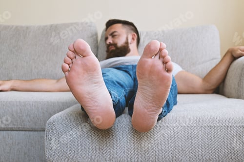 Preview: Bearded barefoot man adult tired sleeping hipster guy on beige sofa, selective focus