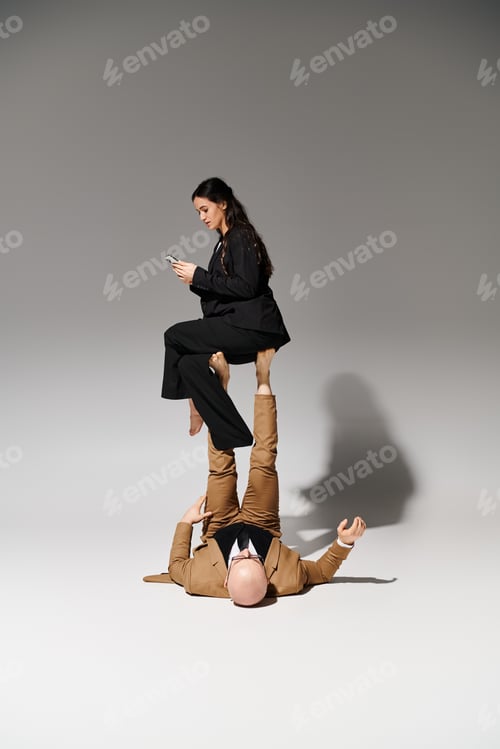 Preview: young businesswoman with smartphone balancing on feet of man in suit on grey, couple of acrobats