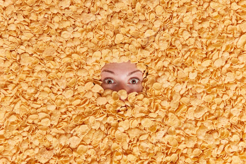 Preview: Shocked young woman buried in cereals has healthy nutrition surriunded by cornflakes hides in food i