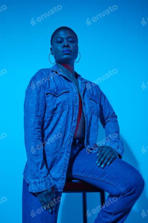 Preview: confident plus size african woman in denim shirt and trousers posing on chair in studio on blue neon