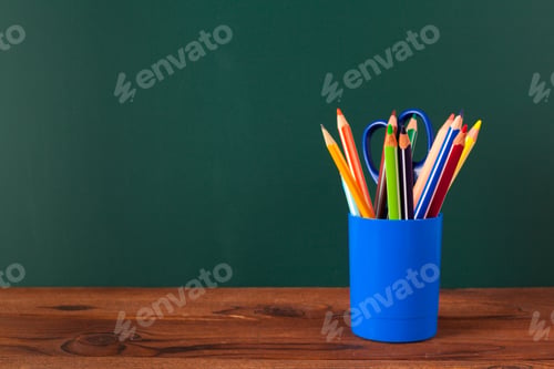 Preview: Colored pencils in a blue cup on a wooden table against a green backdrop