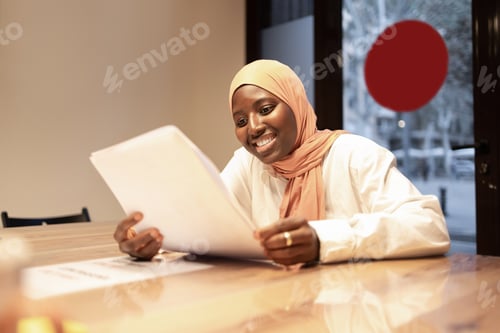 Preview: Smiling muslim businesswoman wearing headscarf sitting at table with papers and reading documents