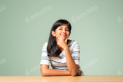 Preview: Indian asian small girl sitting at table against plain green background