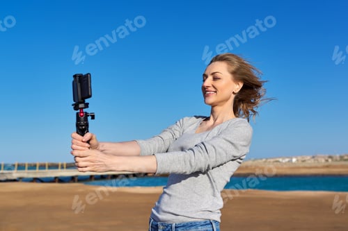 Preview: Middle-aged woman recording video on smartphone with outdoor on seashore