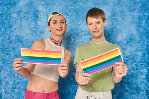Preview: Smiling queer persons in casual clothes holding lgbt flags and looking at camera during pride month