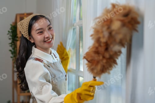 Preview: Young housekeeper wearing yellow gloves is smiling while cleaning white curtains with a feather dust