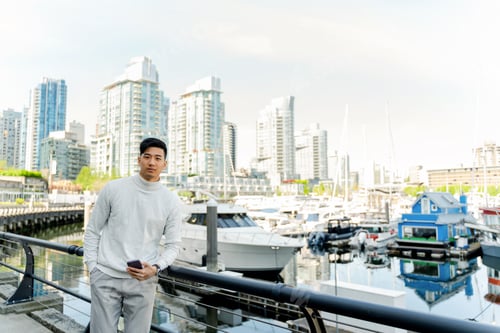 Preview: Сonfident asian man holding mobile phone, standing near expensive yacht in yacht club, looking away