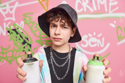 Preview: Stylish serious hipster guy holds two bottles of aerosol spray draws graffiti on street walls being
