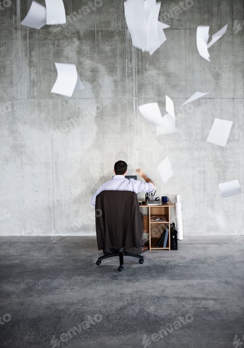 Preview: Caucasian businessman throwing papers in air while sitting at his desk in a large raw space.