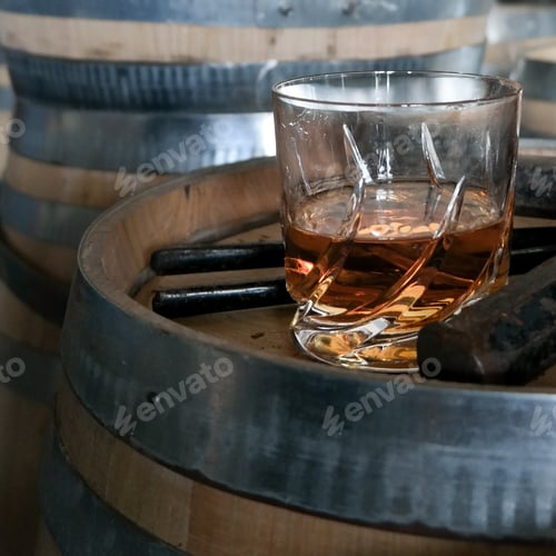 Preview: Glass of whisky cognac or bourbon in ornamental glass next to a vinatge wooden barrel on a rustic wo