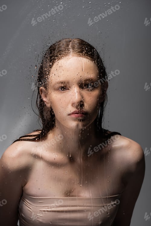 Preview: Young woman looking at camera behind wet glass on grey background