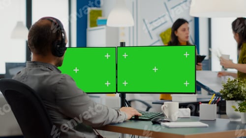 Preview: Employee with headphones working in dual monitror setup with green screen