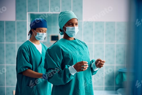 Preview: Surgical nurse helping black female surgeon in getting dressed for operating room in the hospital