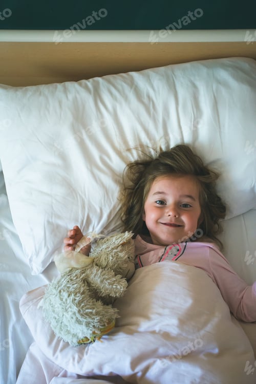 Preview: Little girl lying in a bed with teddy bear at the morning. Having fun in the beginning of the day
