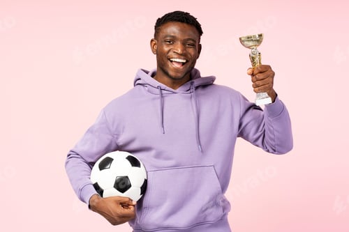 Preview: Portrait of African American athlete celebrating victory, holding a gold cup and soccer ball