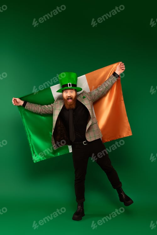 Preview: Full length of excited bearded man in hat holding Irish flag on green background