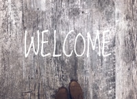 Preview: Top view of reclaimed wood floor with tips of shoes and a WELCOME sign