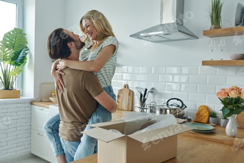 Preview: Young couple embracing and smiling while open box laying on the kitchen counter