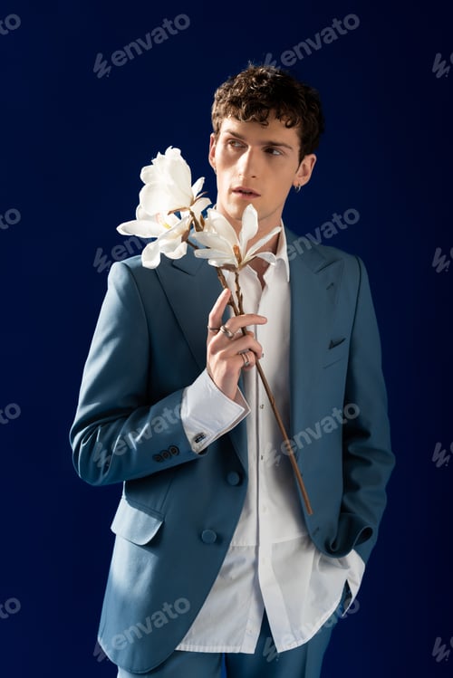 Preview: Fashionable model in suit holding magnolia flowers isolated on navy blue with sunlight