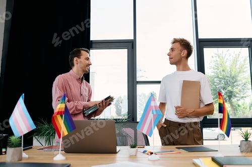 Preview: Happy gay couple discussing creative ideas in a vibrant workspace filled with pride flags