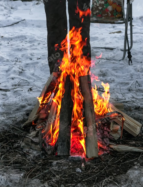 Preview: Female legs by winter campfire on snow at dusk camping in wild, bright red flame of teepee bonfire