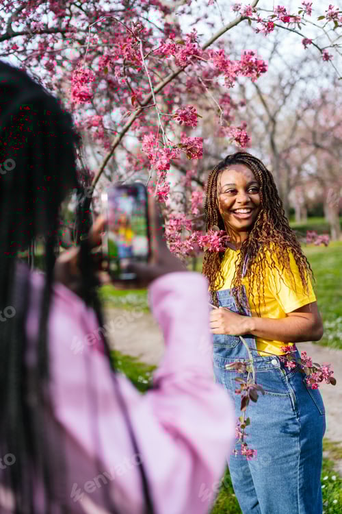 Preview: African woman in yellow shirt smiling for a photo among pink blossoms in a park.