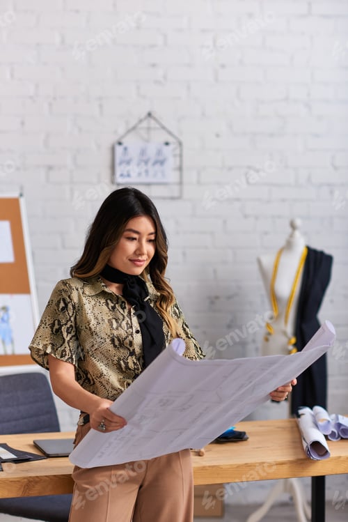 Preview: positive asian designer looking at sewing patterns near work desk in private fashion studio