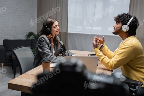 Preview: side view of smiling brunette radio host in headphones and blazer looking at curly indian man