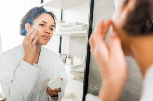Preview: selective focus of african american girl applying moisturizing cream on face in bathroom near mirror