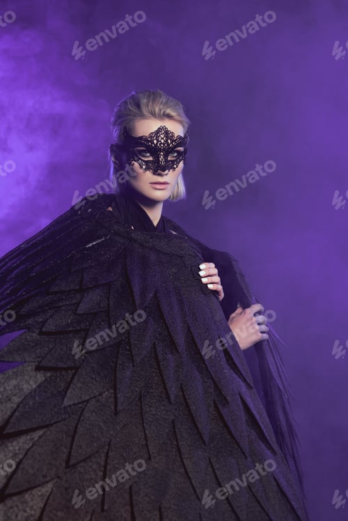 Preview: beautiful woman in lace mask and black angel wings looking at camera and posing on violet background