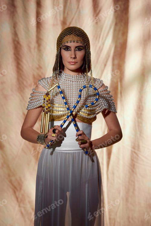 Preview: Woman in egyptian costume and pearl top holding crook and flail while posing on abstract background