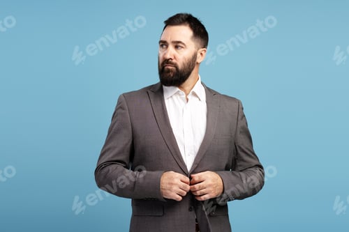 Preview: Confident businessman buttoning his jacket while looking away on blue background