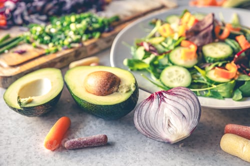 Preview: Avocado halves on the kitchen table with chopped fresh vegetables, close up.