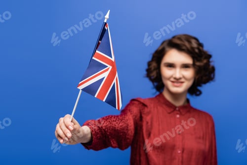 Preview: happy student with curly hair looking at flag of United Kingdom isolated on blue