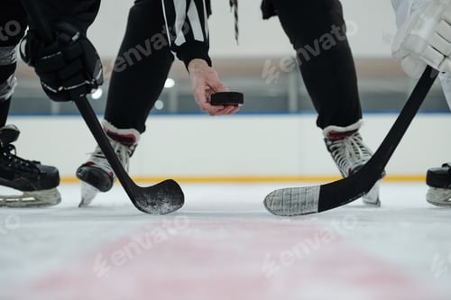 Preview: Hand of referee holding puck over ice rink with two players standing around him
