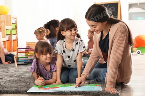 Preview: Kindergarten teacher reading book with cute girls while other children playing together indoors