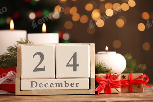 Preview: December 24 - Christmas Eve. Wooden block calendar and decor on table against blurred festive lights