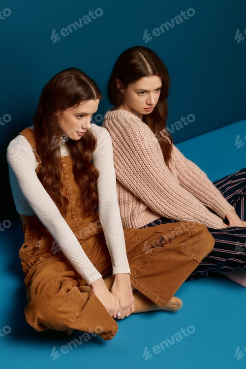 Preview: Beautiful twin sisters share a moment of connection against a vibrant blue backdrop