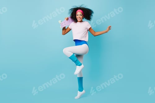 Preview: Good-humoured fitness girl jumping on blue backgorund. Studio shot of glad african woman dancing wi