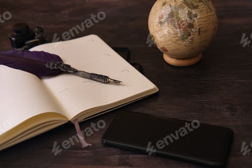 Preview: Closeup shot of a globe, notebook, inkwell, pen, and a cellphone on the wooden table