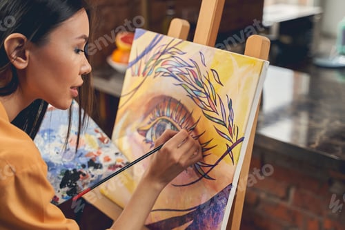 Preview: Charming young woman painting eye on easel at home