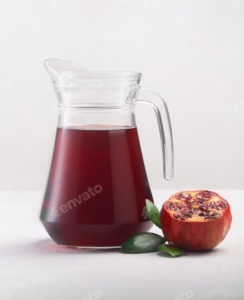Preview: Jug of pomegranate juice with pieces of pomegranate fruits on a white background, close up